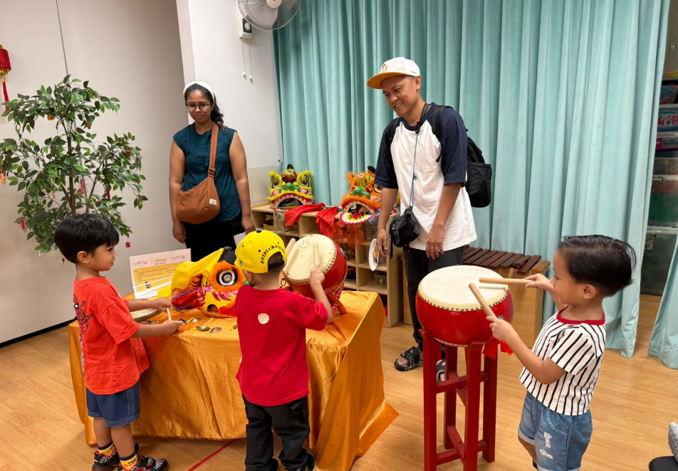 Two adults supervise three children playing drums and instruments beside colorful lion dance heads.